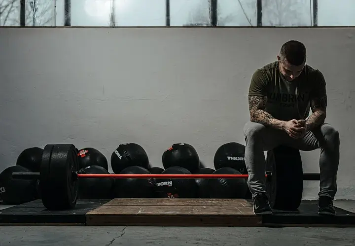 Mężczyzna z tatuażami siedzi na talerzu obciążeniowym, gotowy do treningu crossfit w domu. Obok sztanga i piłki lekarskie.