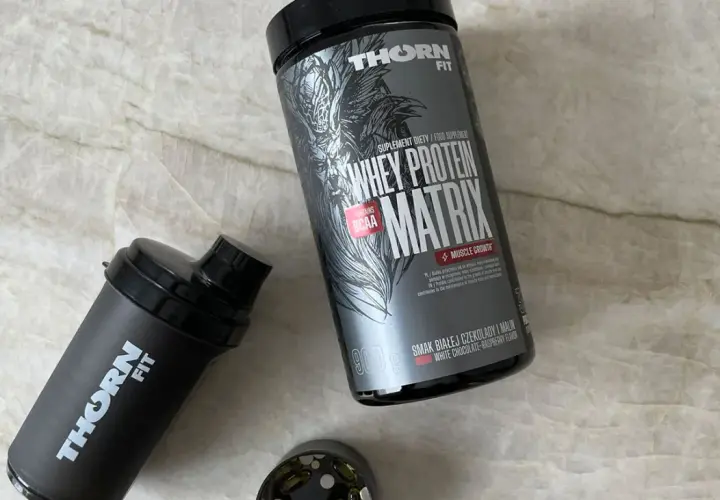 Puszka odżywki białkowej Whey Protein Matrix i czarny shaker marki Thorn Fit na marmurowym tle.