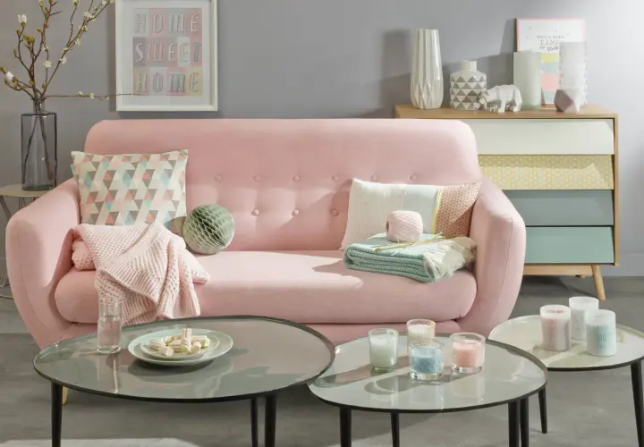 Sofá rosa pastel con cojines y mantas, mesas de centro con velas y malvaviscos, y un mueble de cajones de colores.