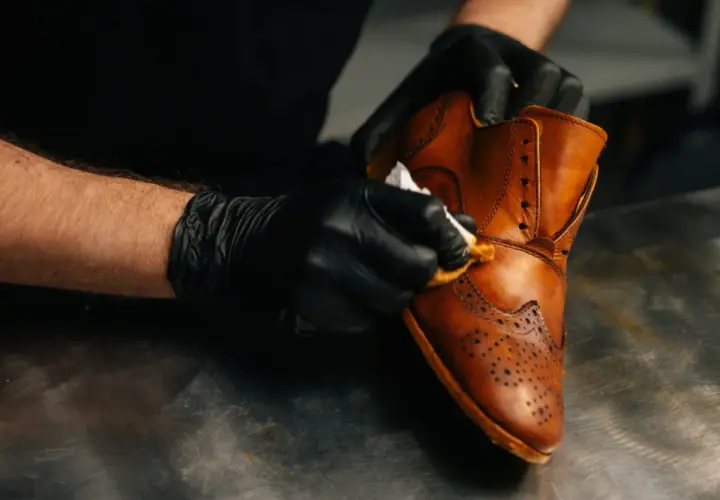 Mains gantées lustrant une chaussure en cuir marron, un soin méticuleux pour un cuir machine à laver qui retrouve son éclat.