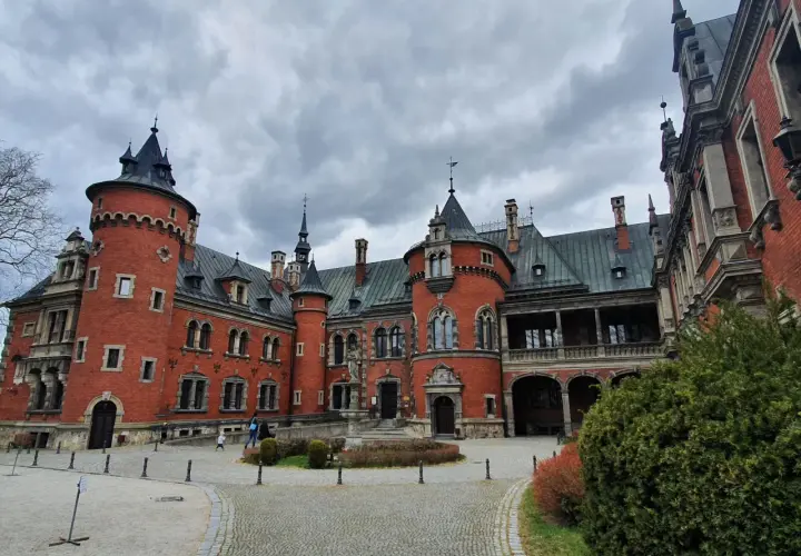 Pławniowice atrakcje: okazały zamek z czerwonej cegły z wieżami i arkadowym gankiem, otoczony brukowanym dziedzińcem i zielenią.