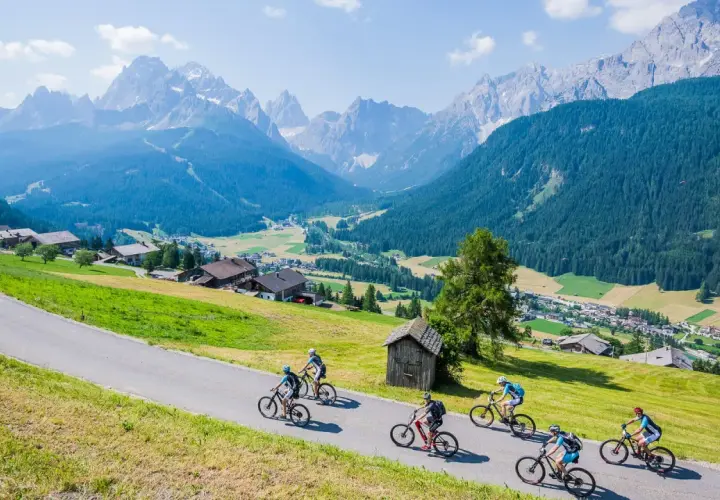 A group enjoys a cycling holiday in the Dolomites, riding down a scenic road with stunning mountain views and a charming village below.