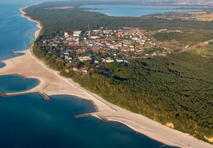 Jarosławiec atrakcje: aerial view of a coastal town with sandy beaches, breakwaters, pine forests, and a lake in the distance.