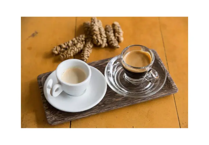 Dwie filiżanki kawy, jedna biała, druga szklana, obok garść ziaren kawy. To kawa z odchodów nietoperza, znana jako Kopi Luwak.