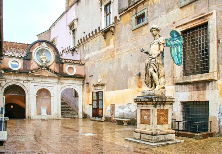 Intérieur du **château Saint-Ange** : une cour pavée et humide, une statue d'ange imposante, une arche menant à un escalier et des fenêtres grillagées.