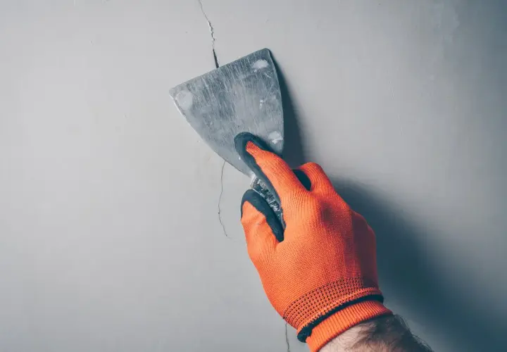 Ręka w pomarańczowej rękawicy wypełnia głębokie pęknięcia w ścianie szpachlą za pomocą kielni.
