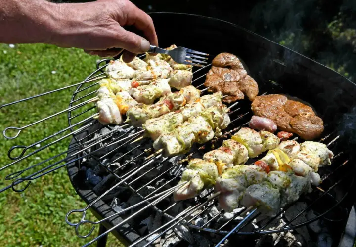 Une main vérifie des brochettes de poulet et des steaks sur un barbecue rustique.