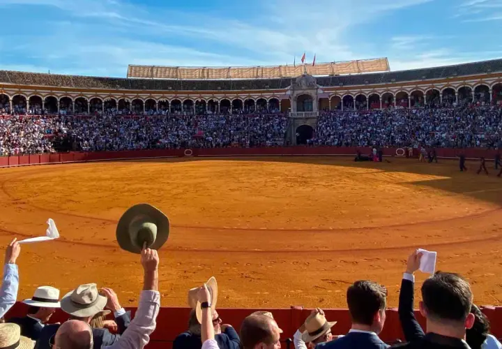Espectadores en una **plaça de toros** levantan pañuelos y sombreros ante la arena dorada y el público en las gradas.