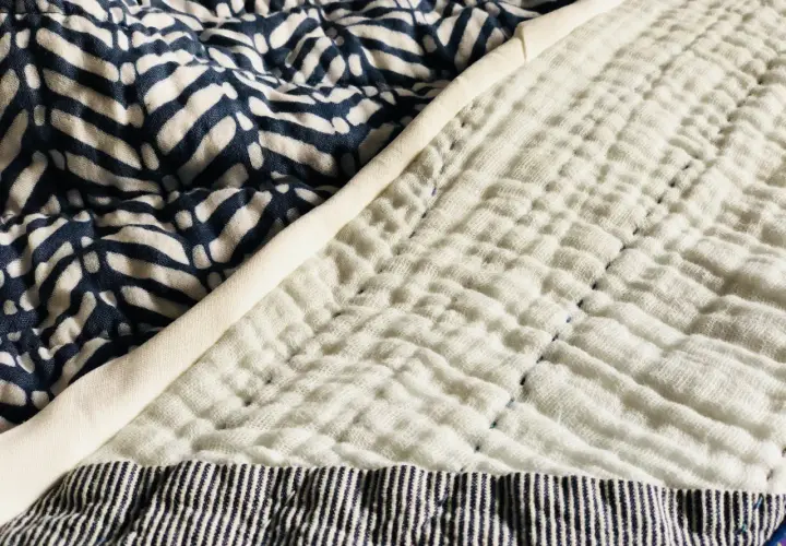 A close-up of a quilt shows a patterned blue fabric next to a textured white double gauze fabric, stitched with blue thread.