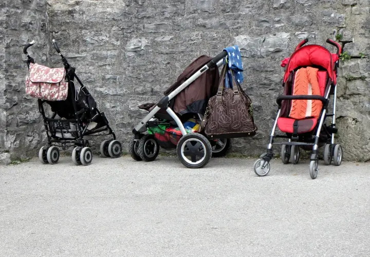 Trzy puste wózki dziecięce stoją przy kamiennej ścianie. Jeden ma torbę w kwiaty, drugi brązową torbę i niebieską chustę, a trzeci pomarańczowy kocyk.