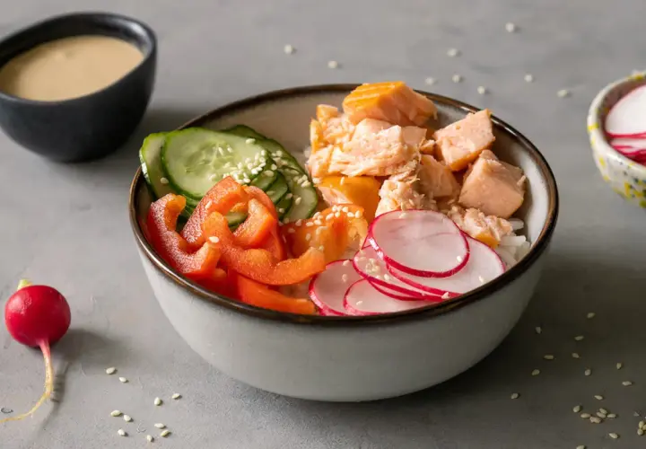 Kolorowa sałatka do sushi z łososiem, ryżem, ogórkiem, papryką i rzodkiewką. Obok miseczka z sosem.