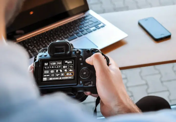 Ręce trzymają aparat, na ekranie ustawienia manualne. Nauka fotografii w praktyce, obok laptop i telefon.