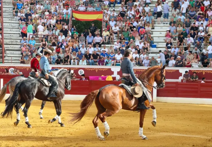 Dos jinetes, uno con chaquetilla roja y otro con azul, montan caballos en una plaza de toros. Bernardino Piriz, con sombrero, observa la escena.
