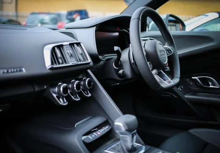 Interno di un'auto sportiva con volante a sinistra. Il cruscotto è scuro, con bocchette d'aria cromate e un display digitale. La leva del cambio è in primo piano. Perfetta per esplorare paesi con guida a sinistra.