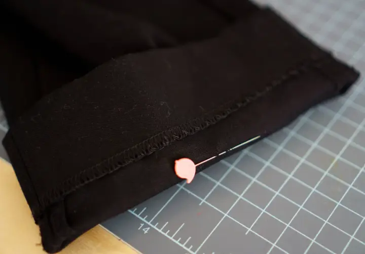 Close-up of black pants hem, pinned and ready for an invisible stitch. A pink pin secures the fabric on a cutting mat.