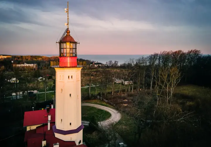 Najstarsza latarnia morska w Polsce, wysoka, kremowa wieża z czerwonymi akcentami, góruje nad krajobrazem o zachodzie słońca.