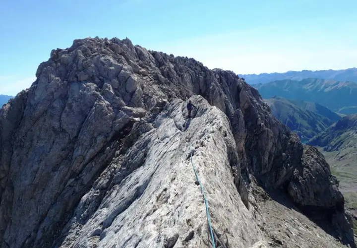 Alpinista en la cresta rocosa del Pico de Vallibierna, con un arnés y cuerda azul. Paisaje montañoso al fondo.