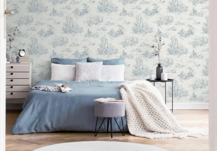 Chambre paisible avec un papier peint au motif jouy bleu et blanc, un lit bleu, des coussins blancs et une couverture en maille.