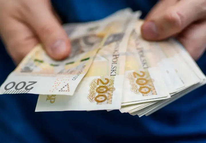 Dłonie trzymają plik banknotów 200 zł. Pytanie brzmi, po jakim czasie od zarejestrowania wypłacany jest zasiłek dla bezrobotnych.