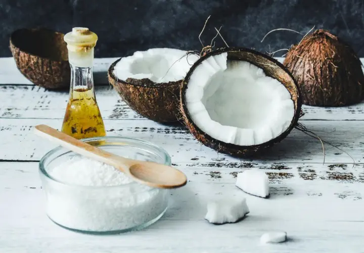 Coupé en deux, un coco frais et sa chair blanche côtoient une petite fiole d'huile dorée. Un bol de noix de coco râpée et une cuillère en bois suggèrent l'utilisation de l'huile de coco pour des recettes gourmandes ou des soins.