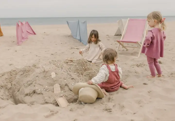 Dzieci budują zamek z piasku, ciesząc się beztroskimi zabawami na plaży. W tle widać leżaki i parawany.