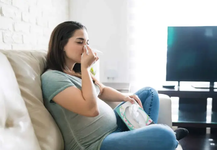Femme enceinte s'essuyant le nez avec un mouchoir, inquiète d'un rhume enceinte danger pour bébé. Elle est assise sur un canapé, une boîte de mouchoirs à portée de main.