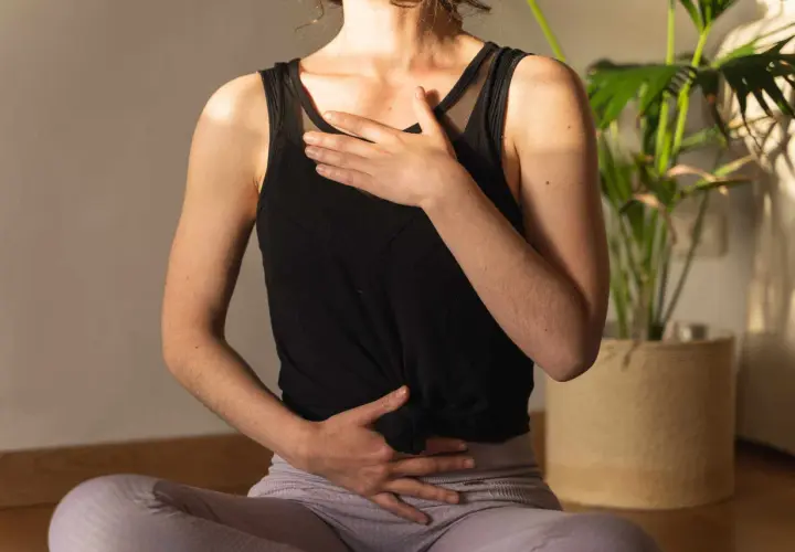 Une femme assise en tailleur, mains sur le ventre et la poitrine, pratique la respiration profonde. Une plante verte est visible en arrière-plan.