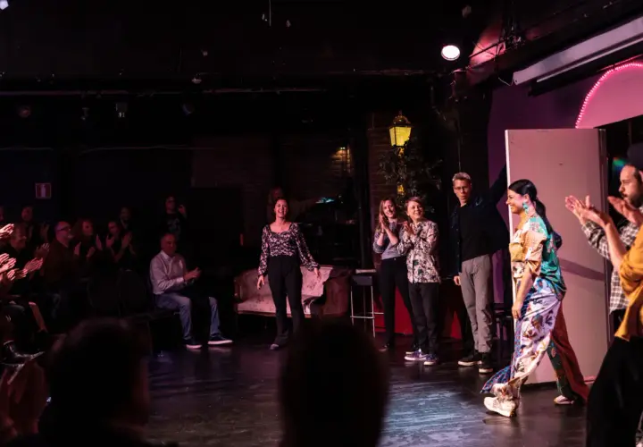 Publik applåderar en föreställning av improvisationsteater i Stockholm. Skådespelare på scenen tar emot hyllningar.