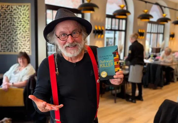 Klaus-Peter Wolf präsentiert "Ostfriesenkiller", Band 1 seiner Krimireihe. Ein Mann mit Hut und roten Hosenträgern hält das Buch hoch.