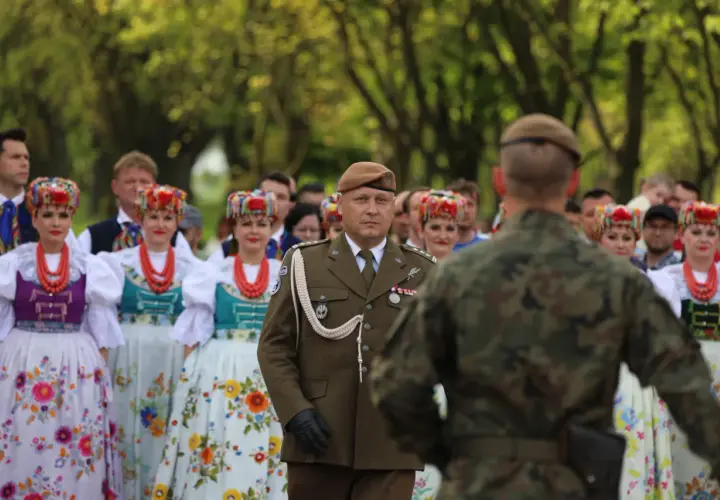 Żołnierz w brązowym capstrzyku i mundurze stoi przed grupą kobiet w tradycyjnych, kolorowych strojach ludowych.