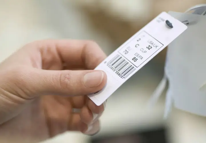 Main d'une personne tenant une étiquette de vêtement avec un code-barres et des tailles.