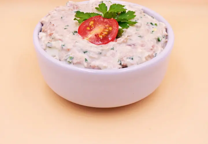 Pyszna pasta z tuńczyka i jajka, udekorowana pomidorkiem i natką pietruszki, w białej miseczce.