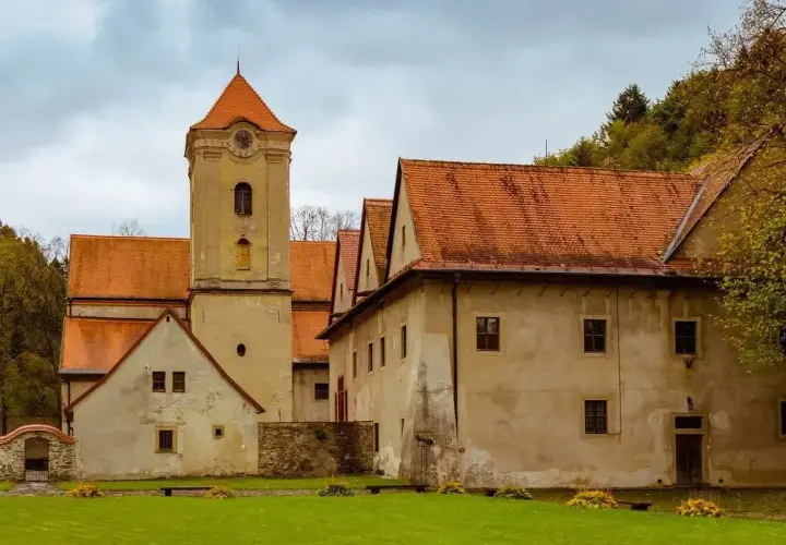 Czerwony klasztor zaprasza na zwiedzanie. Zabytkowa wieża i budynki z pomarańczowymi dachami otoczone zielenią.