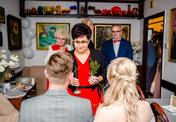 Kobieta w czerwonej sukience udziela nowoczesne błogosławieństwo parze młodej, trzymając gałązkę.