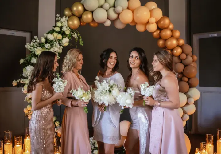 Cinq femmes souriantes, entourées de fleurs et de ballons, se préparent pour un mariage. Elles tiennent des bouquets, prêtes pour la cérémonie.