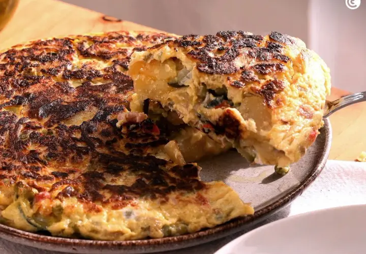 Una porción de tortilla paisana, dorada y jugosa, se levanta con un tenedor, revelando patatas, pimientos y huevo.