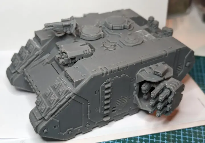 Szary model Land Raider Redeemer, gotowy do malowania, z działkiem na dachu i wyrzutniami flamerów po bokach.