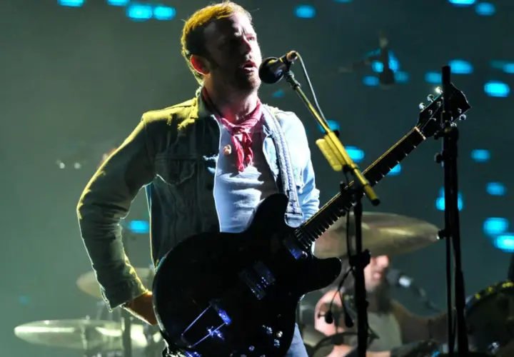 Wokalista Kings of Leon Polska na koncercie, gra na gitarze elektrycznej, w dżinsowej kurtce i czerwonej chuście.