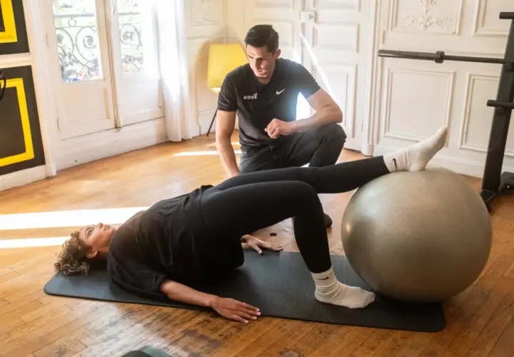 Un homme observe une femme faire un pont sur un tapis, les pieds sur un ballon de gym. Un exercice pour le bienfait d'étirement et de renforcement.