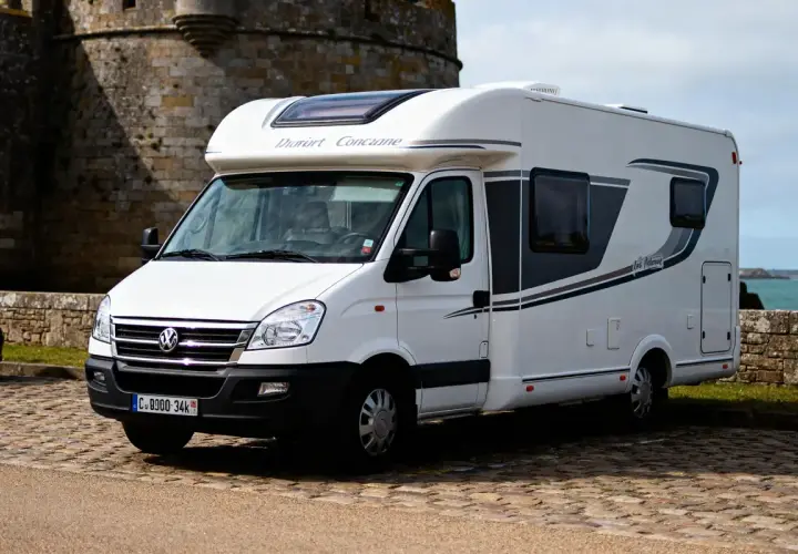 Un camping-car blanc "Dauphin Concarneau" stationné sur un sol pavé, avec un mur de pierre et la mer en arrière-plan. Un spot camping car Bretagne idéal pour explorer la côte.