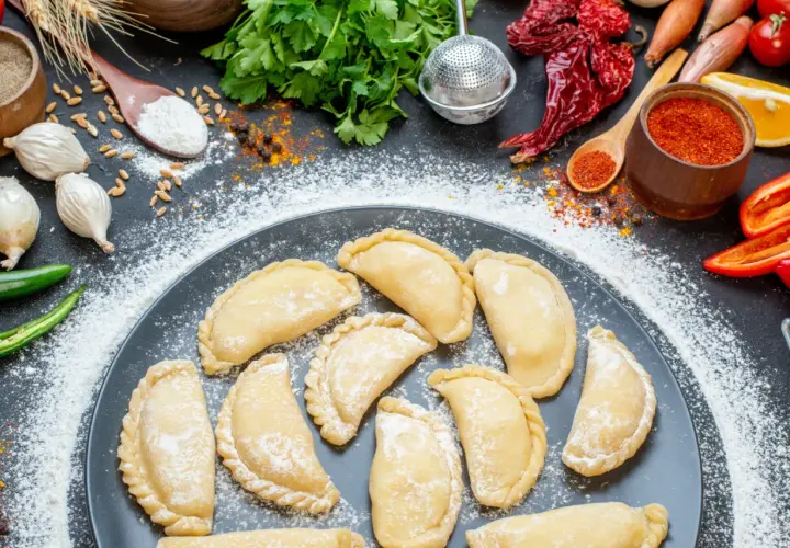 Gotowi, by nauczyć się, jak zrobić pierogi? Na talerzu surowe pierożki, obok mąka, cebula, papryczki chili i przyprawy.