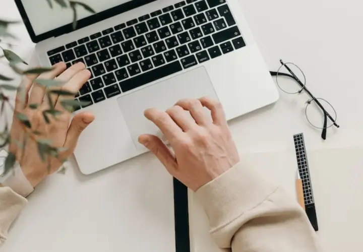 Ręce piszą na klawiaturze, obok leżą okulary i notatnik. Idealny tani laptop na studia, by zacząć naukę.