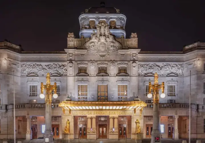 Kungliga Dramatiska Teatern i Stockholm, upplyst på natten. En storslagen byggnad med detaljerad fasad och gyllene utsmyckningar.