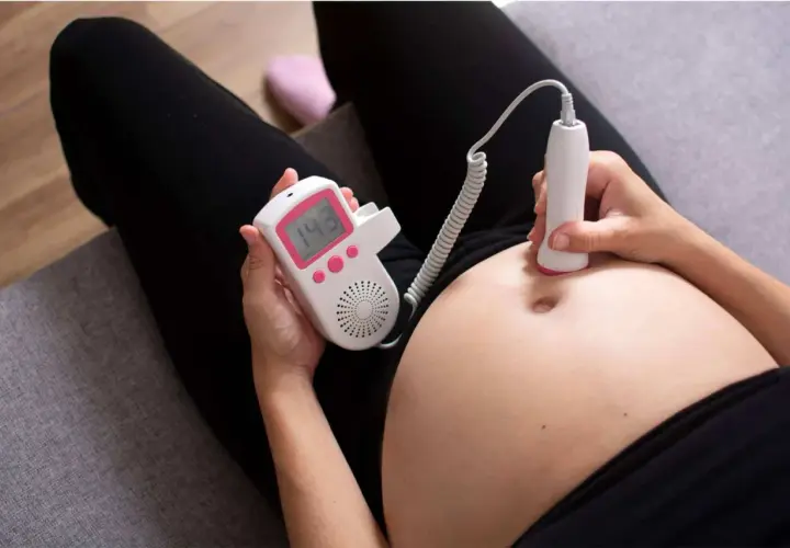 Una donna incinta usa un doppler fetale per ascoltare il battito del feto toccando la pancia. Il display mostra 143 bpm.