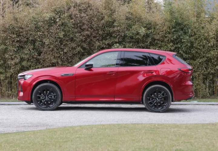 Czerwona Mazda CX-60 3.3 diesel z czarnymi felgami stoi na żwirowej drodze, za nią gęsty żywopłot.