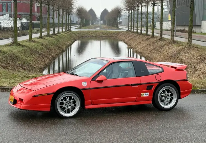 Roter Pontiac Fiero GT mit Felgen steht auf nasser Straße neben einem Kanal.