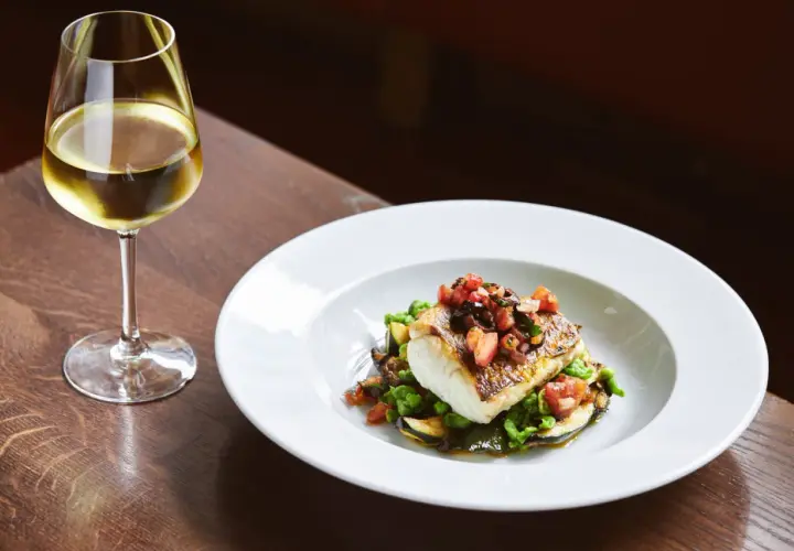 Un verre de vin blanc accompagne un poisson en sauce, servi sur un lit de légumes verts et de courgettes.
