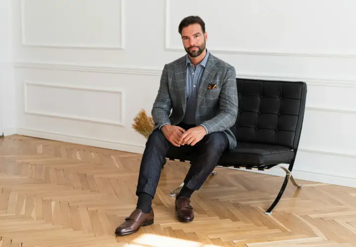 Elegancki mężczyzna w szarej marynarce i granatowych spodniach, z brązowymi butami do jeansów i marynarki, siedzi na czarnym fotelu.