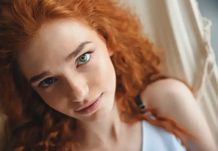 Primer plano de una joven con cabello pelirrojo natural y ojos claros, recostada en una hamaca.