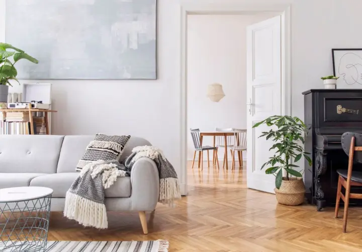 Un salón moderno con sofá gris, manta, cojín, mesa de café y piano negro. La luz blanca ilumina el espacio, creando una atmósfera acogedora.